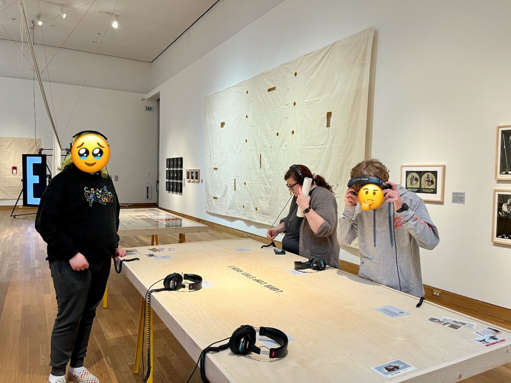 Two teenagers and their mother surrounding a table, listening to audio recording on headphones. They are in an art gallery with art pieces on the walls behind them.