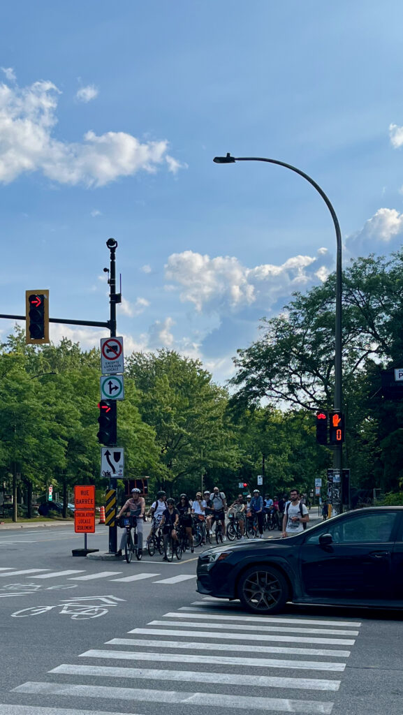 The image shows a city intersection with a group of cyclists waiting at a red light on a dedicated bike lane. The intersection has various traffic lights, including one specifically for cyclists. There are trees and greenery in the background, and the sky is clear with a few clouds. A pedestrian is also waiting at the crosswalk, while a dark-colored car is partially visible in the foreground, about to make a turn. A sign in French reads "Rue Barrée," indicating a street closure ahead, with another sign allowing exceptions for local traffic.