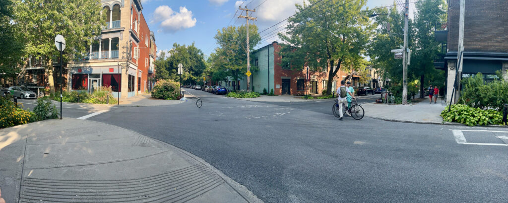 The image shows a quiet, urban street corner with low-rise buildings, trees, and greenery. A cyclist is walking their bike across the intersection, while another person follows. The buildings feature a mix of architectural styles, including brick and stone facades. The street is lined with parked cars, and there are a few pedestrians walking on the sidewalks. The intersection has crosswalks, and the atmosphere appears calm and residential, with a mix of greenery and urban elements.