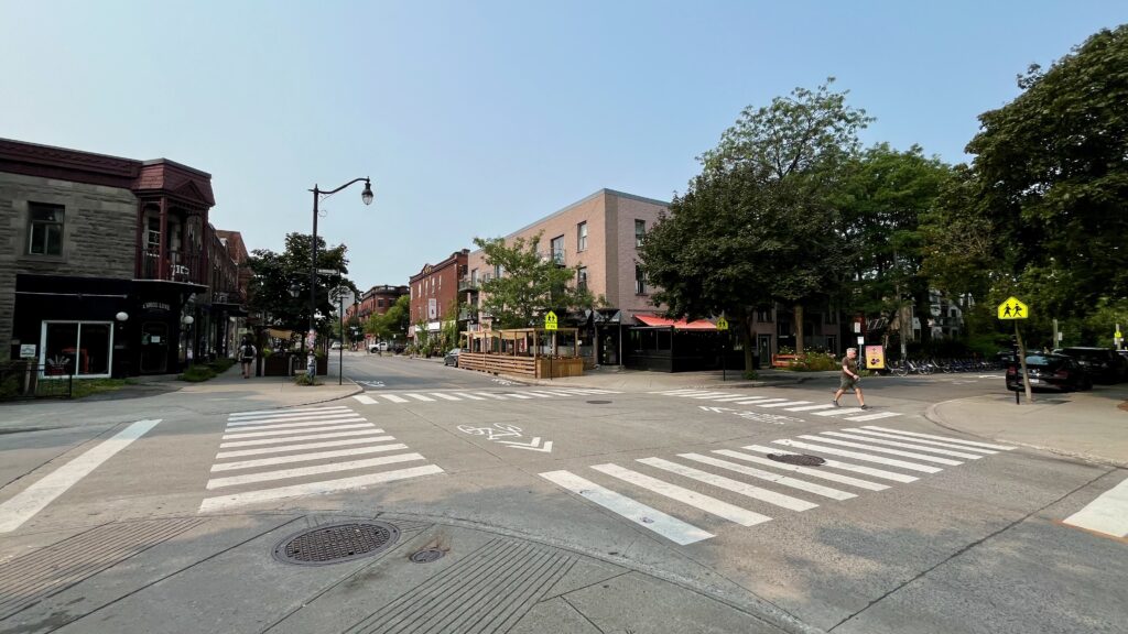 A bustling street in Montreal,
 Quebec. There are several buildings on either side of the street, including a red brick building with a stone facade. Pedestrians are crossing the street at a crosswalk, and there are parked cars along the curb.