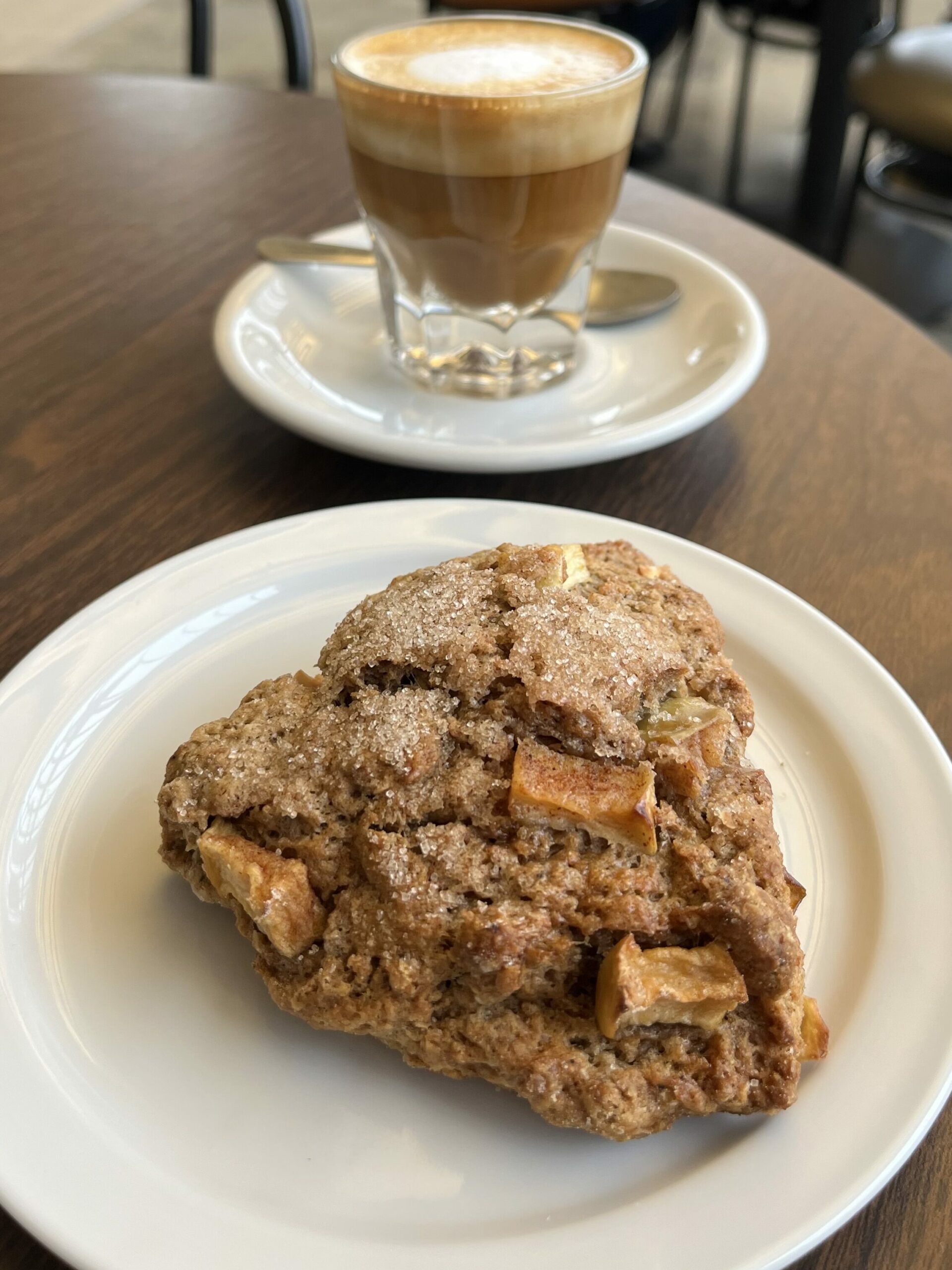 A rustic-looking scone with visible chunks of apple baked into it, dusted with sugar crystals on top, served on a white plate. Behind it is a cortado, served in a clear glass on a white saucer.