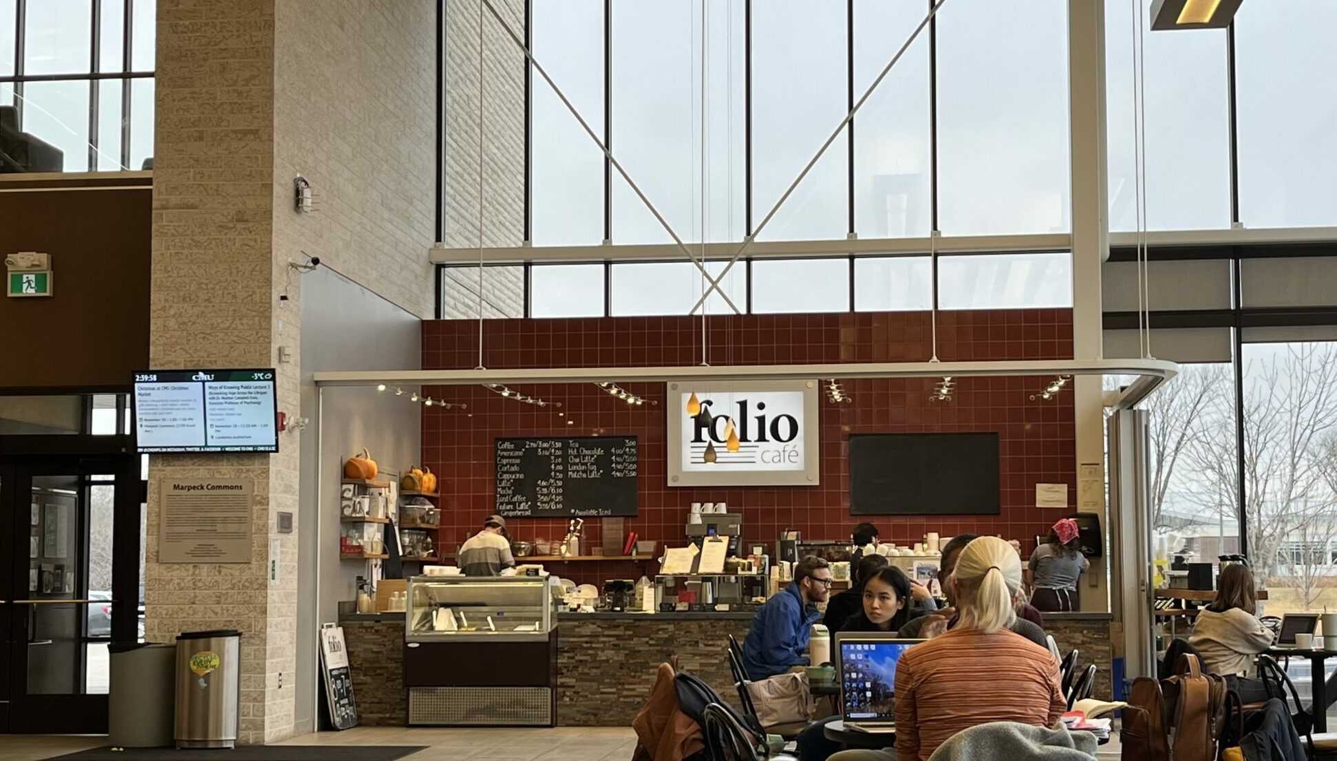 The café has red tile walls, floor-to-ceiling windows, and a counter with a display case. Several students are seated at tables with laptops. The space features high ceilings and modern architecture with exposed structural elements. A green exit sign is visible near the entrance.