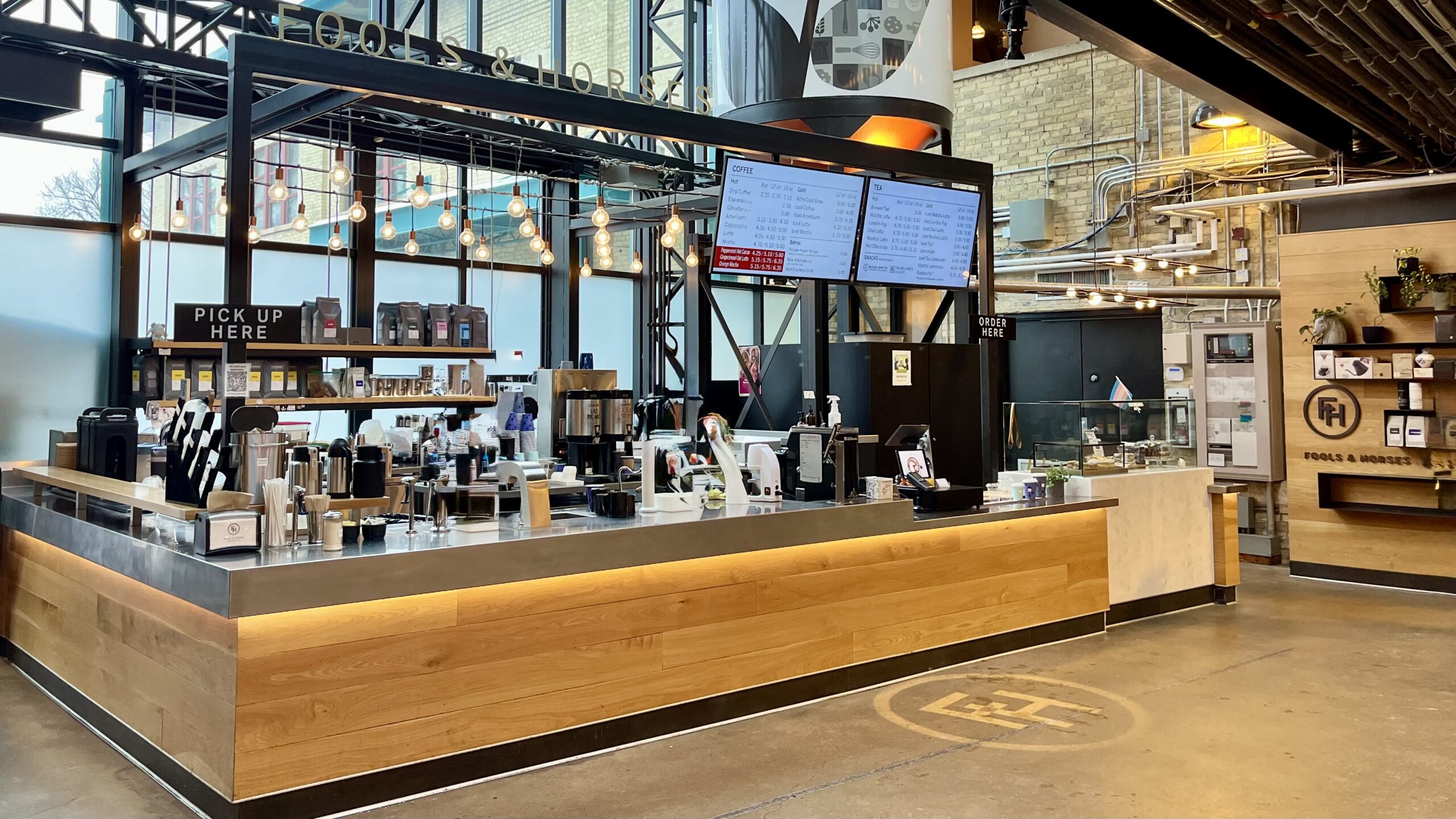 Interior of a modern coffee shop with 'FOOD & DRINK' signage visible above. The service counter features a wooden base with stainless steel top, decorated with hanging Edison bulb lighting. Digital menu boards are mounted above. The counter is well-equipped with coffee-making equipment, and there's a 'PICK UP HERE' sign on the left. The space has an industrial design with exposed ceiling elements and large windows. A FH symbol is visible on the floor.