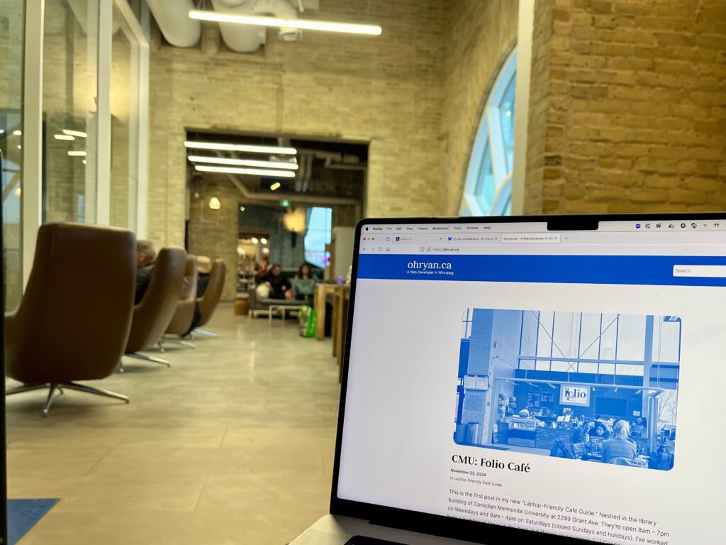 A laptop screen showing a website 'ohryan.ca' with an article about CMU's Folio Café, featuring a blue-tinted image of the café. The laptop is positioned in what appears to be the actual café, with brown modern swivel chairs and exposed brick walls visible in the background.