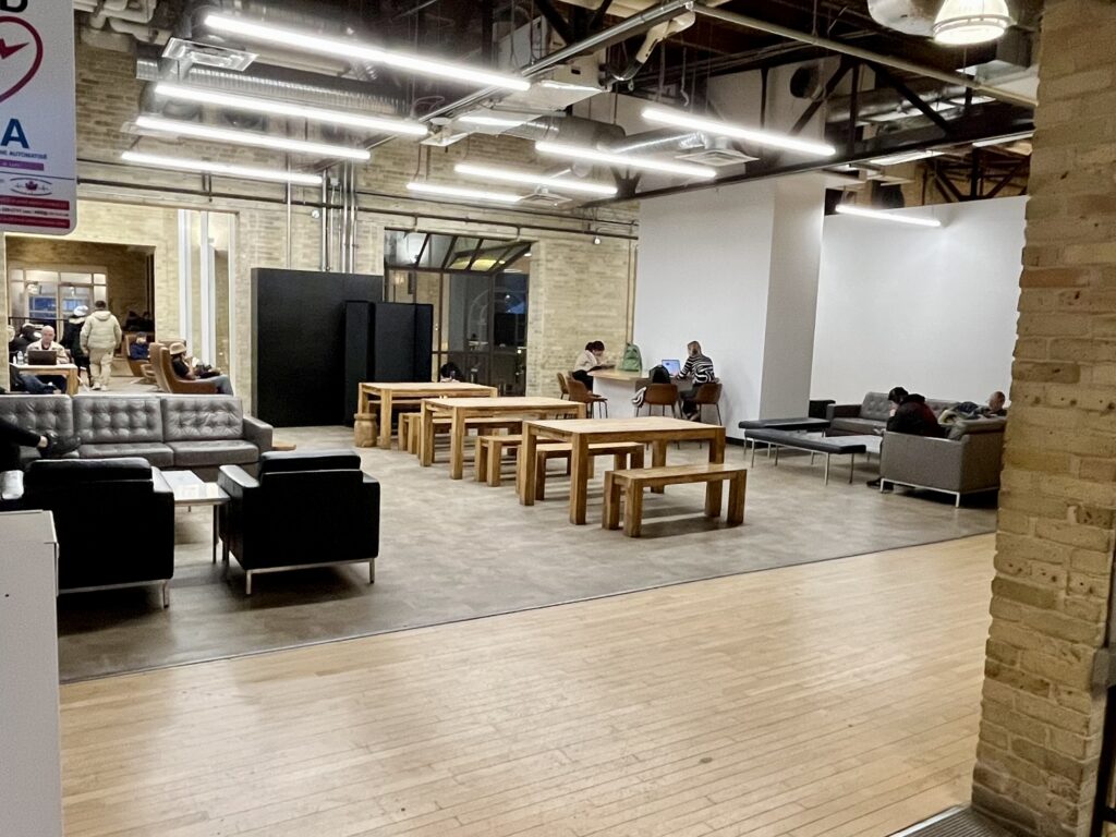 A modern study or lounge space with industrial-style exposed ceiling and lighting. The room features a mix of seating options including black leather armchairs, grey sofas, and wooden communal tables with benches. The floor transitions from wood to concrete, and the space has exposed brick walls. A few people are seated throughout the space studying or working.