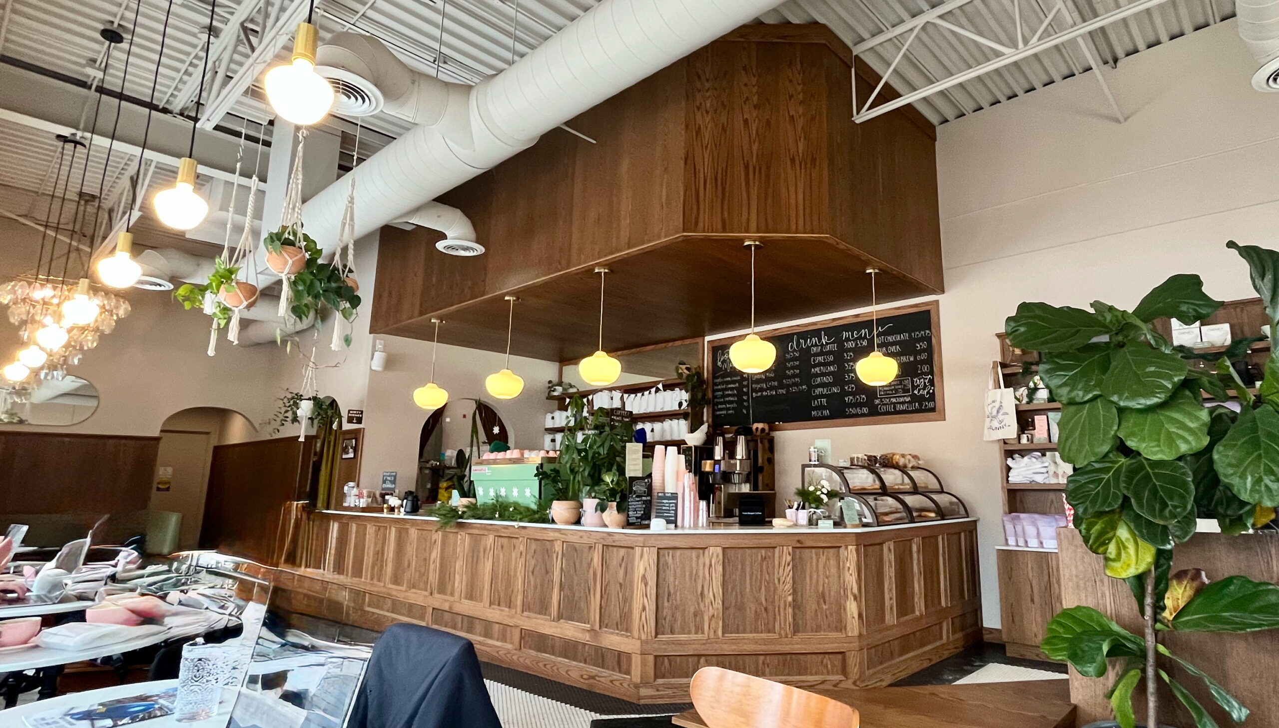 Interior of a cozy café with a wooden paneled counter and display case. Features yellow pendant lights, hanging macramé planters with greenery, and a chalkboard menu on the wall. The ceiling has exposed white ductwork, and there are large monstera plants decorating the space. The overall design combines natural wood elements with industrial touches.