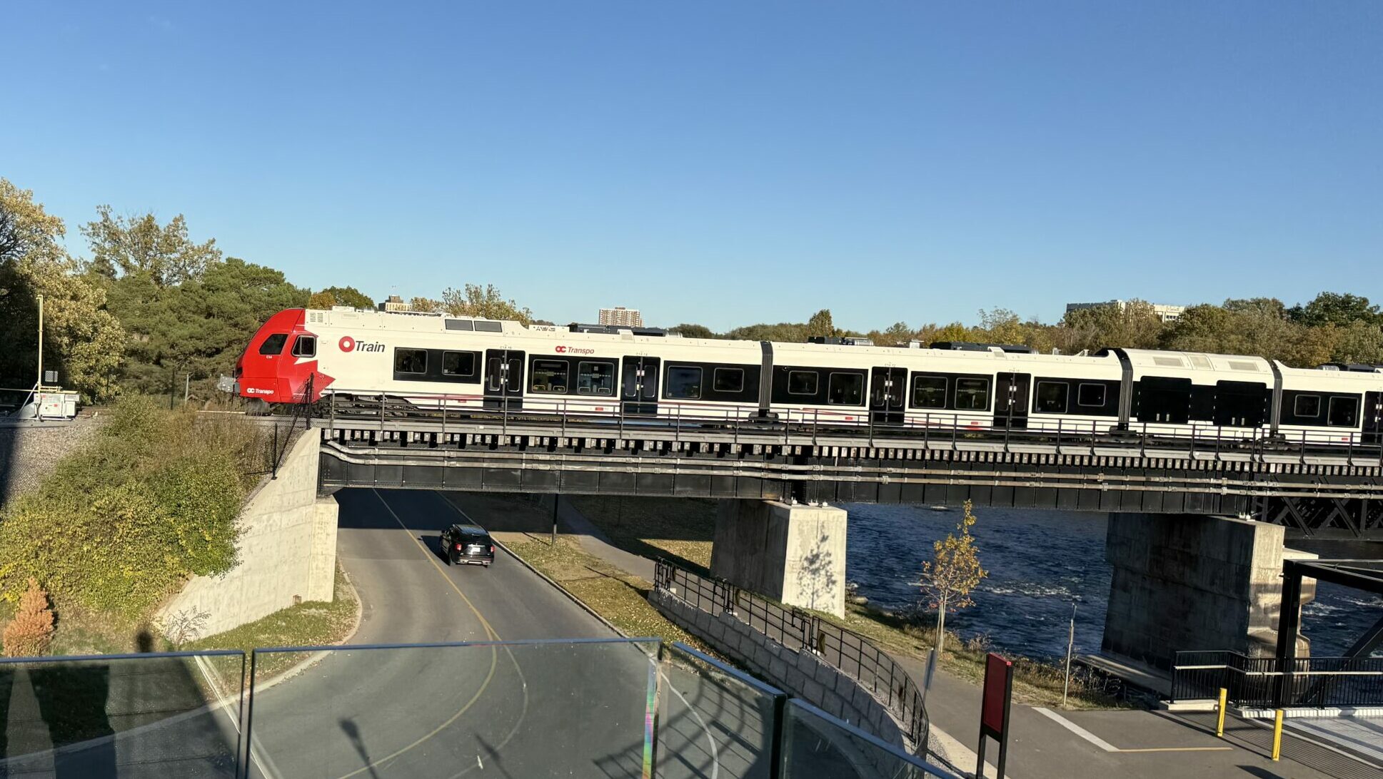 A red and white OC Transpo O-Train travels across a steel bridge over a river in Ottawa on a clear autumn afternoon. Below the bridge, a car drives along a curved road and a bike path runs parallel to the river. Trees with early fall foliage surround the scene, and the photo is taken from a higher glass-railed viewpoint.
